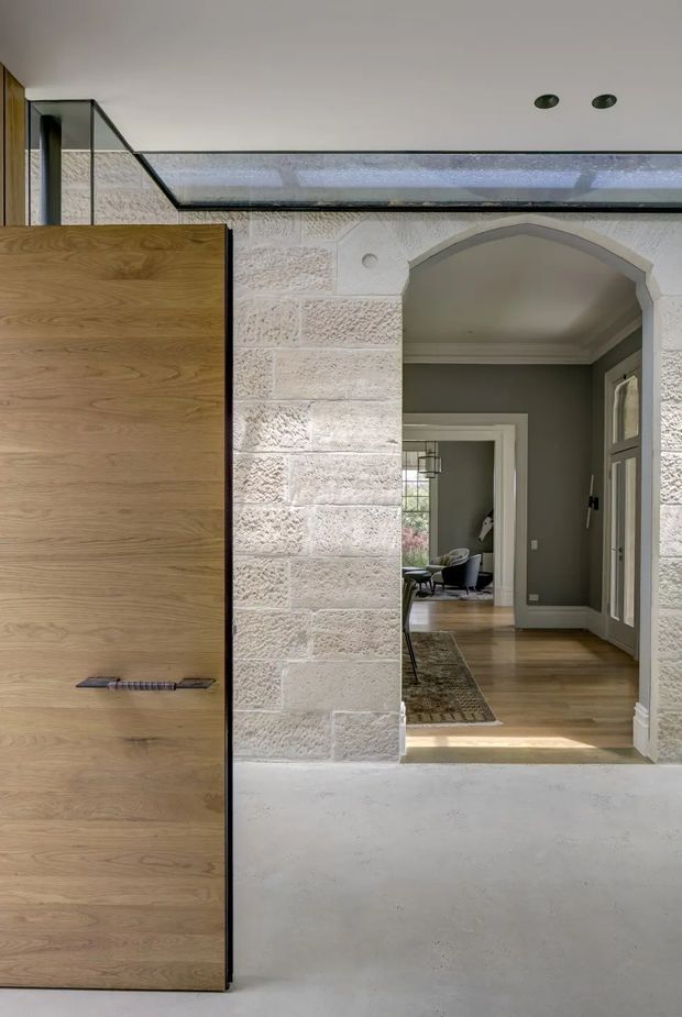Interior view of a refined transitional hallway or entry passage with a large natural timber built-in door or cabinet panel in the foreground, pale textured masonry block walls, a broad glazed clerestory above, and an arched opening leading into a furnished living area. The space combines warm timber flooring and grey-green walls with crisp white trim and restrained recessed lighting.