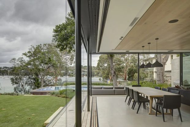 Contemporary open-plan dining and living area opening onto a landscaped waterfront-facing lawn through extensive floor-to-ceiling glazing. The space features a long dining table with dark upholstered chairs, a grey sofa, sculptural pendant lights, a pale timber-lined ceiling, dark slender window frames, and polished light flooring. A pool and waterway are visible beyond the lawn, with mature trees surrounding the property.