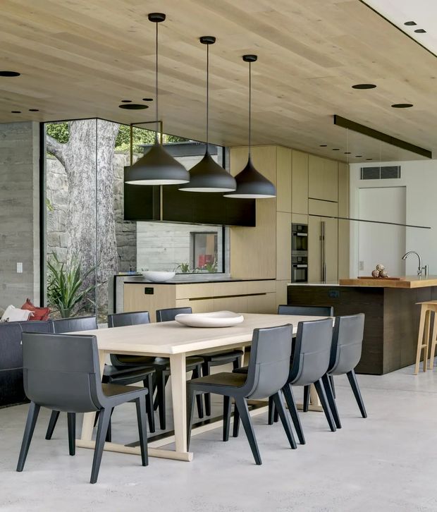 A refined open-plan kitchen and dining area with a large light timber dining table surrounded by dark upholstered chairs. The kitchen features pale timber flat-panel cabinetry, a dark stone or composite island benchtop, integrated appliances, a timber-lined ceiling, concrete walls and floor, and a dramatic cluster of dark pendant lights. Large corner glazing opens the room toward a planted outdoor area, while a lounge is partially visible to the left.