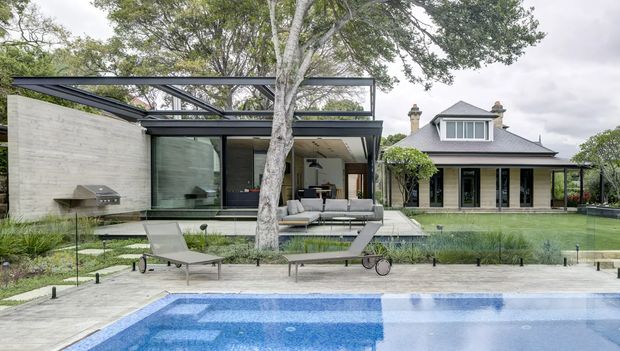 A landscaped residential courtyard viewed from beside a swimming pool, featuring a contemporary flat-roof pavilion with expansive glazing and concrete walls alongside a more traditional two-storey gabled house. The space includes a paved pool terrace, sun loungers, lawn, ornamental grasses, mature trees, and outdoor seating. The architecture contrasts modern and traditional forms within a wooded suburban setting.