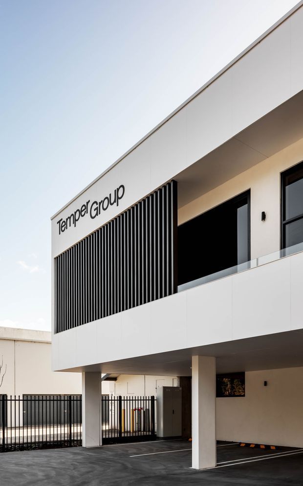 A contemporary two-storey commercial building exterior with a clean white facade, deep overhangs, black-framed glazing, a glass-balustraded balcony, and a prominent vertical black louvre screen. The building is elevated over a marked covered parking area, with perimeter fencing and signage reading “TemperGroup.”