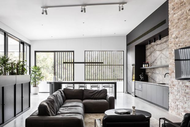 Open-plan contemporary living room with a large dark sectional sofa facing a low coffee table and wall-mounted television. The space includes a sleek dark kitchen and bar area, black-framed glazed doors and windows, a pale polished floor, white walls, exposed light brickwork, a dramatic marble-look splashback, indoor plants, and track lighting.