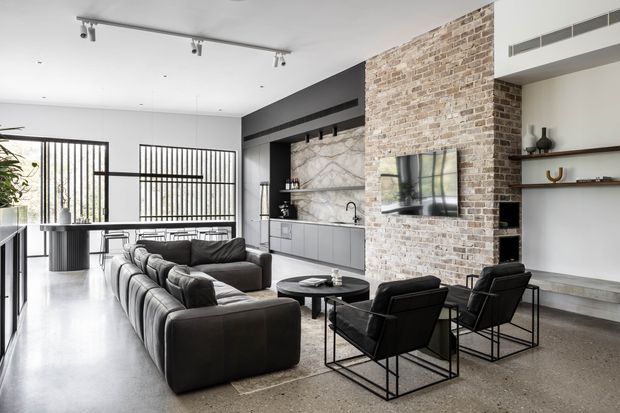 Open-plan contemporary living area with a large dark sectional sofa, black lounge chairs, round coffee table, and integrated kitchen cabinetry. A tall exposed light-brick feature wall incorporates a television and fireplace, while a dark marble-look splashback, black-framed glazing, polished concrete-style flooring, and open shelving create an industrial-modern interior.