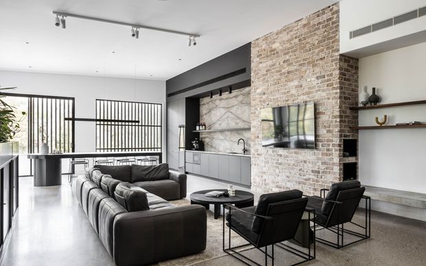 Open-plan contemporary living area with a large dark sectional sofa, black lounge chairs and round coffee table arranged around a media wall. A substantial exposed light-brick feature wall divides the living space from a sleek grey kitchen, with marble-look backsplash, integrated cabinetry and a peninsula. Black-framed full-height glazing with vertical screening brings in daylight, while open shelving and minimalist track lighting complete the refined industrial-modern interior.