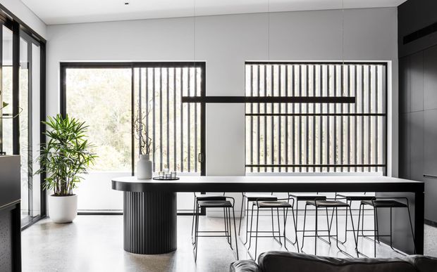 Contemporary open-plan kitchen and dining area with a long black island or dining counter, slim black bar stools, dark integrated cabinetry, large windows with vertical screening, and a sculptural black pendant light. The neutral grey and white palette is softened by a potted indoor plant, while the polished concrete floor and crisp built-in joinery create a refined minimalist feel.