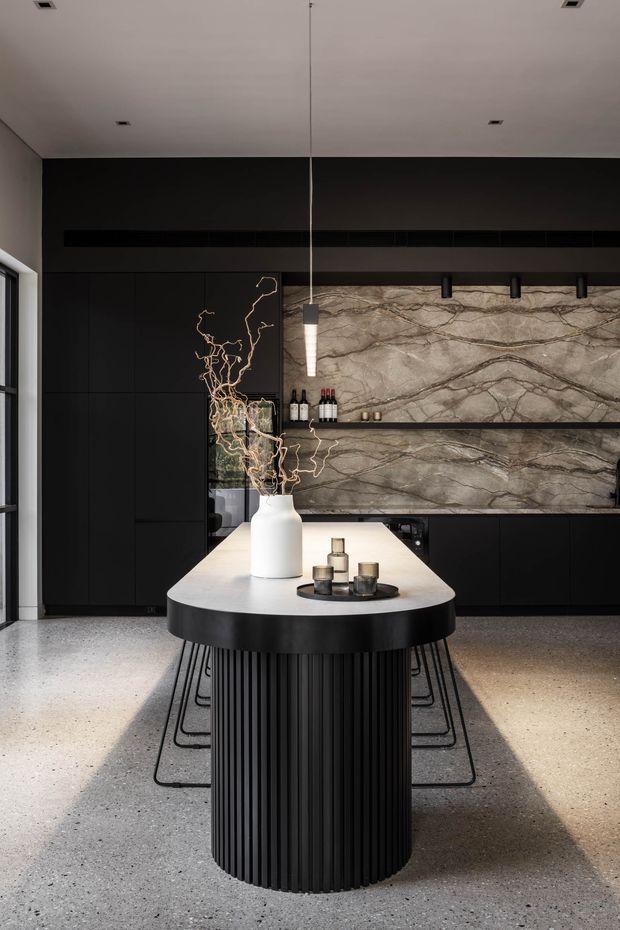 A sophisticated contemporary kitchen with a long oval island featuring a fluted black base, pale stone benchtop, and slim black bar stools. The room has matte-black full-height cabinetry, a dramatic veined stone backsplash and shelf, dark integrated ventilation, a polished concrete or terrazzo floor, and minimalist pendant and recessed lighting. A large window on the left brings in natural light.
