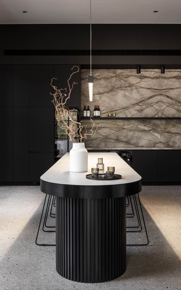 A dramatic, refined contemporary kitchen centred on a large rounded island with a fluted black base and pale stone benchtop. Matte-black full-height cabinetry contrasts with a veined stone splashback and open shelf, while a linear pendant and recessed ceiling lights provide focused illumination. Bar stools surround the island, and a sculptural white vase with branches forms the centrepiece.