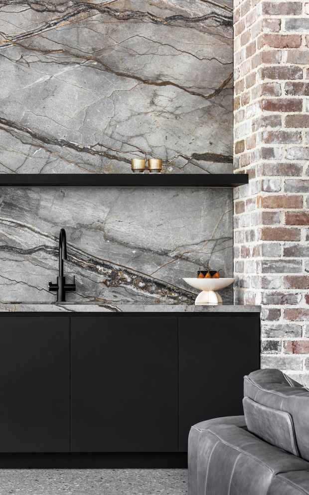 A dramatic contemporary kitchen or entertaining space featuring a large grey, heavily veined natural-stone splashback and wall finish, paired with matte black cabinetry, black benchtops, and a black tap. An exposed white-and-brown brick wall forms the right side of the composition, while a grey upholstered sofa is visible in the foreground, suggesting an open-plan connection to a living area. Warm metallic candle holders and a small decorative bowl add subtle accents.