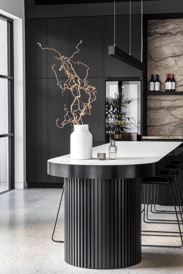 A sophisticated contemporary kitchen and dining area featuring a sculptural black island with a fluted cylindrical base and pale stone benchtop. Matte black full-height cabinetry forms the backdrop, paired with a veined stone feature wall and integrated wine storage. Linear black pendant lighting hangs above the island, while dark bar stools and a large window complete the refined monochrome scheme.