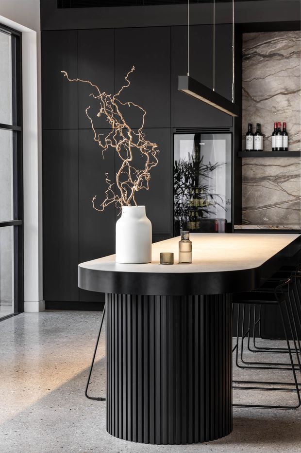 A dramatic contemporary kitchen and dining space featuring a sculptural black fluted island with a pale stone benchtop, dark full-height cabinetry, a marble-look feature wall with open wine shelving, integrated refrigeration, and a linear pendant light. A large window and polished concrete-style floor add natural light and texture.