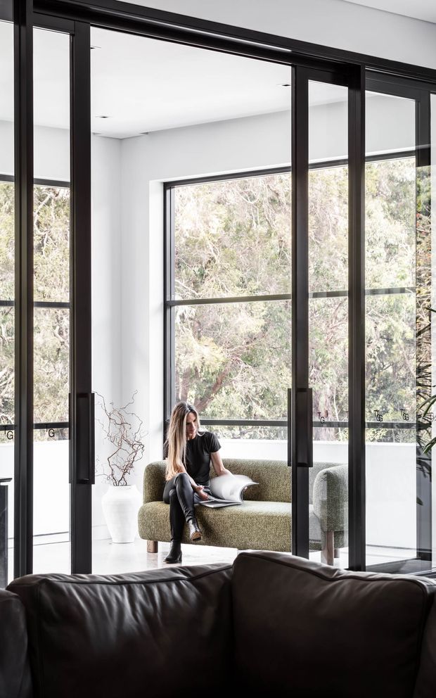 Bright contemporary living area with full-height black-framed glazed doors and windows overlooking trees. A person sits on a textured green upholstered sofa in a small glazed sitting nook, while a dark leather sofa occupies the foreground. White walls, recessed ceiling lighting, and strong indoor–outdoor visual connection create a clean, refined interior.