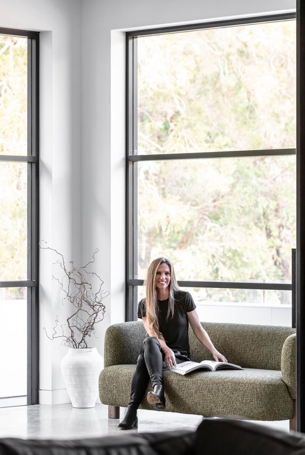 Bright contemporary living room with a woman seated on a textured olive-green sofa, large black-framed floor-to-ceiling windows, white walls, and a polished light floor. A simple white ceramic vase with sculptural branches adds a restrained decorative element, while greenery is visible outside.