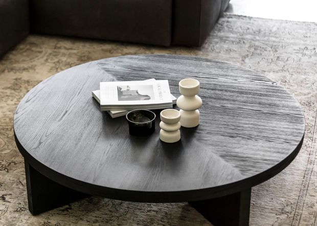 A round, low-profile dark wood coffee table with a broad oval top and chunky triangular support legs, styled with stacked books and small ceramic vessels. It sits on a large patterned rug in front of a dark sofa, creating a refined, monochromatic living-room vignette.