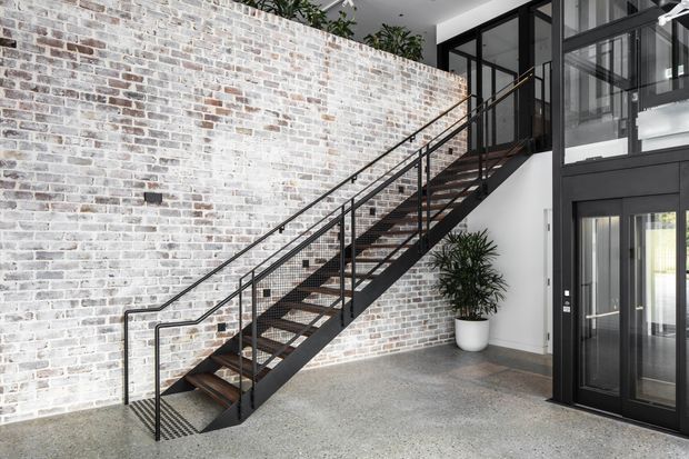 Contemporary interior entry space featuring a dramatic open-riser black metal staircase with timber treads, a large whitewashed exposed-brick feature wall, black-framed glazing, and a glazed lift or entry enclosure. The polished grey floor and potted greenery add a refined industrial character.