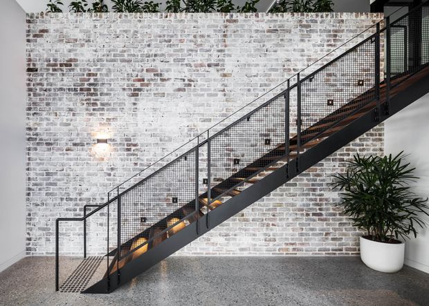 A contemporary interior stairway with a straight open-riser staircase, black metal stringer and mesh balustrades, timber treads, and warm integrated lighting. The staircase runs diagonally across a whitewashed exposed brick feature wall, with a polished grey concrete floor and potted greenery.