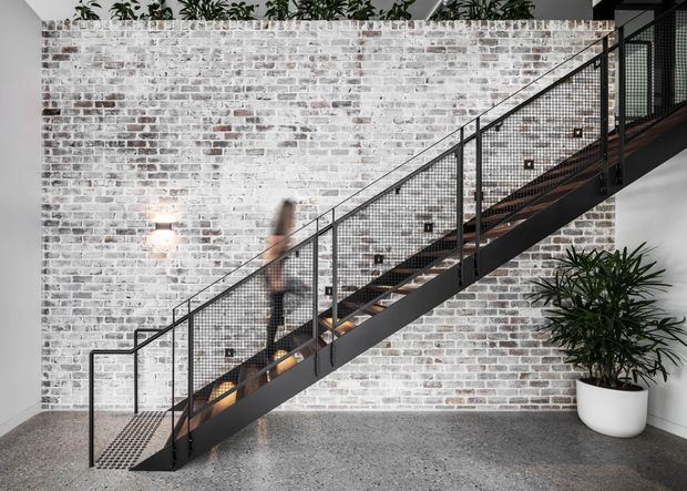 Interior view of a modern industrial stairway with a dark metal stringer, mesh balustrade, and timber stair treads set against a whitewashed exposed brick wall. The space has a polished grey concrete floor, potted greenery, and contemporary wall lighting.