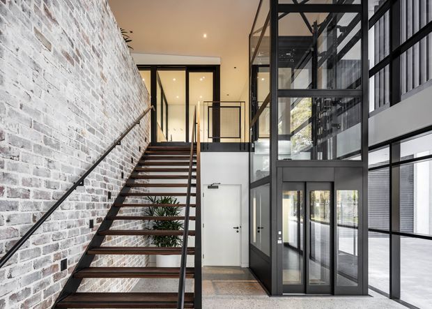 A bright contemporary multi-level interior featuring an open industrial-style stairway beside a white-painted exposed brick wall. Black metal stair framing, balustrades, and extensive black-framed glazing surround a white door and polished concrete-like floor, with a planted area visible beneath the stairs.