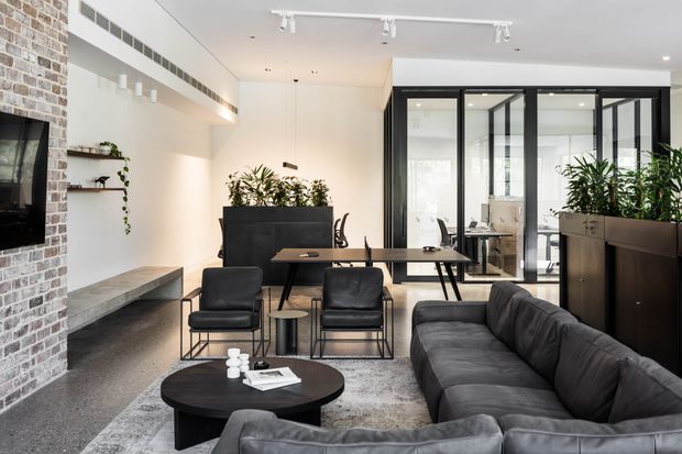 A bright, contemporary open-plan living room with a large charcoal sectional sofa, black round coffee table, leather lounge chairs, and a dining or work table. The room features a white ceiling and walls, polished concrete flooring, a full-height exposed brick feature wall with a wall-mounted television and built-in bench, black-framed glazed partitions, indoor plants, and streamlined black cabinetry. Track lighting and linear ventilation details contribute to the modern industrial character.