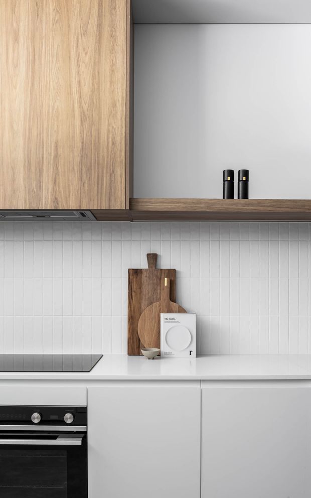A close-up view of a contemporary kitchen featuring flat-panel light timber overhead cabinetry, dark timber trim and open shelving, matte white lower cabinetry, a white benchtop, and a vertically ribbed white tile splashback. A black cooktop and built-in oven occupy the left side, while black tapware and timber cutting boards provide contrast.