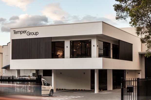 Exterior view of a contemporary two-storey commercial building with a crisp white rectangular facade, deep overhangs, dark framed glazing, vertical black screening, and a covered ground-level parking or entry area. The TemperGroup sign is visible on the upper facade, with a gated forecourt and adjacent urban landscaping.