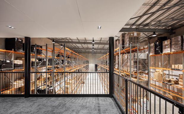 Interior view of a large industrial warehouse or distribution facility from an elevated mezzanine walkway, with extensive pallet racking, boxed inventory, black metal balustrades, exposed structural services, and a mix of suspended industrial lighting and smooth ceiling panels.