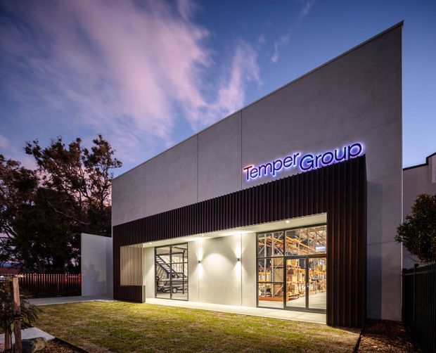 Exterior dusk photograph of a contemporary commercial warehouse or showroom building. The facade combines smooth pale rendered or concrete panels with dark vertical metal screening, large glazed entry and display windows, and illuminated “TemperGroup” signage. Shelving and stored materials are visible through the glazing, with landscaped lawn and trees in front.
