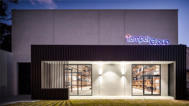 Night-time exterior view of a contemporary commercial building with a smooth grey facade, dark vertical slatted screening around the entrance, illuminated glazed display or retail windows, and a neon-style “Temper Group” sign.