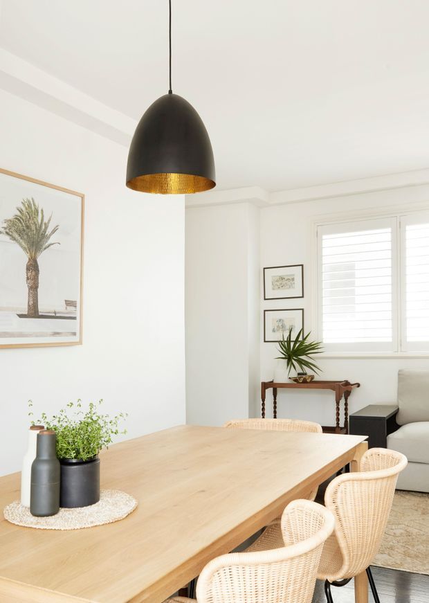 Bright, refined dining area with a large light timber dining table and woven rattan dining chairs. The room features white walls and ceiling with simple decorative trim, a large black pendant light with a textured gold-toned interior, white plantation shutters, framed botanical artwork, and a small timber console with indoor plants. A portion of an adjacent living area is visible at right.