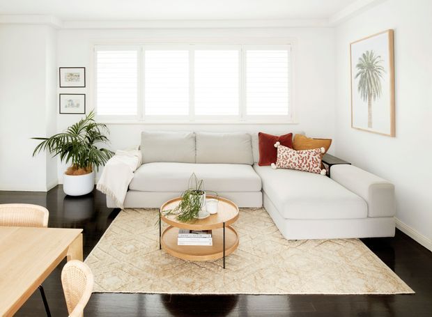 Bright, refined living room with a white sectional sofa, layered patterned cushions, a round wood-and-glass coffee table, large pale rug, indoor palm, and dark timber flooring. White plantation shutters provide strong natural light, while framed botanical artwork and light furnishings create a calm contemporary look.