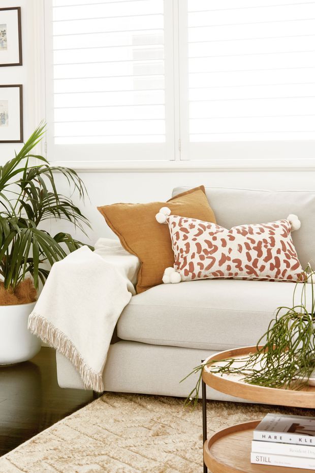 Bright, cozy living room vignette featuring a cream upholstered sofa, rust and patterned throw pillows, a fringed cream blanket, large indoor palms, white plantation shutters, framed artwork, a plush neutral rug, and round wood-and-metal nesting coffee tables with books.