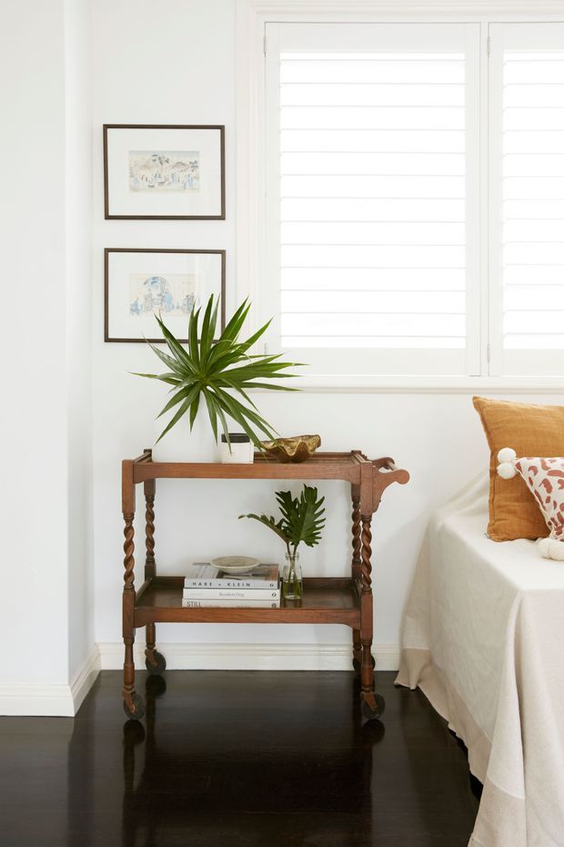 Bright, softly styled bedroom corner featuring a dark polished timber floor, white walls and plantation shutters, a traditional carved timber console table, framed artwork, tropical foliage, and the edge of a bed with warm rust-toned cushions and a patterned accent pillow.