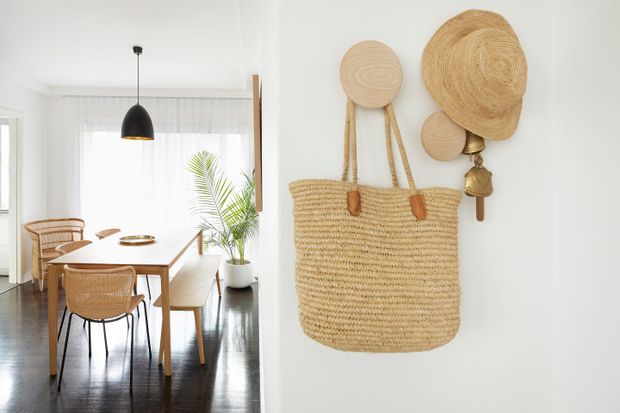 Bright dining area with a light timber dining table, woven cane chairs, a bench seat, and a black pendant light. A white wall in the foreground displays natural woven baskets and a large woven tote, while a potted palm adds greenery near the window. Dark timber flooring contrasts with the predominantly white and natural-fibre palette.