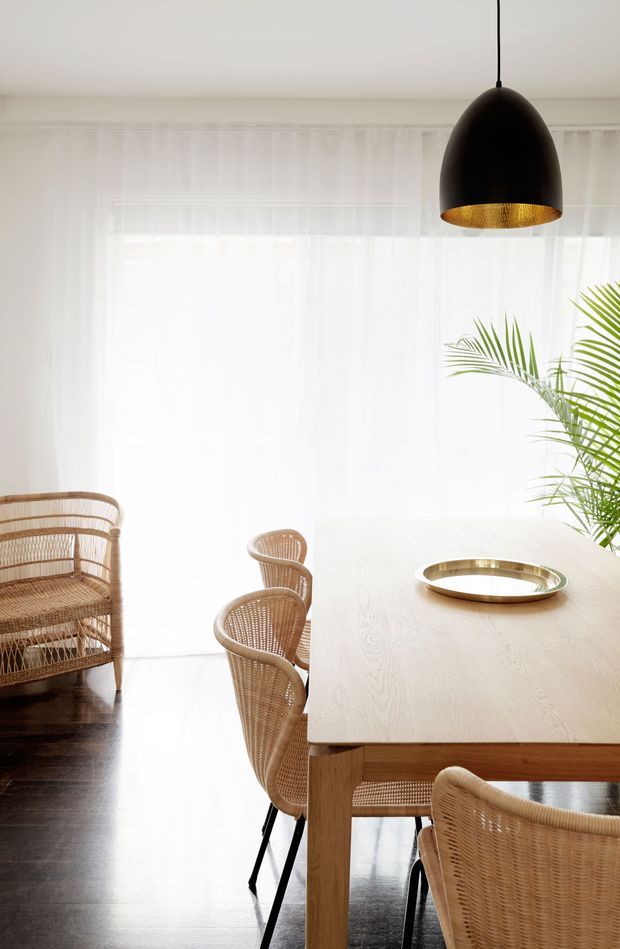 Bright dining area with a large light timber dining table, woven rattan dining chairs and armchair, dark timber flooring, sheer white curtains, a black pendant light with a gold-toned interior, and a tall palm plant. The space has an airy, natural, relaxed contemporary look.