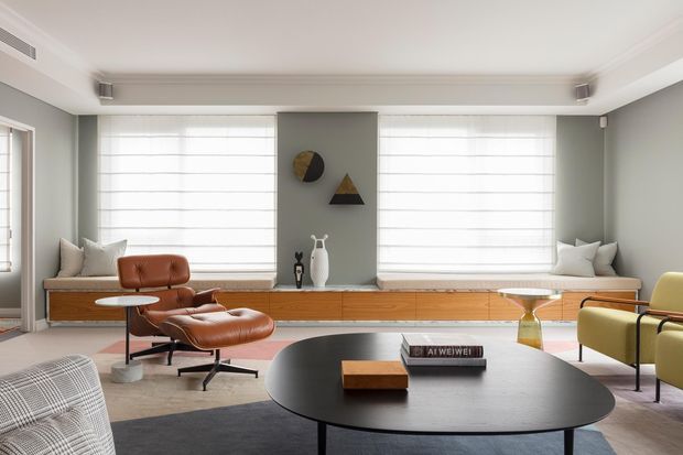 Bright, refined mid-century-inspired living room with pale grey walls, broad windows fitted with white Roman blinds, and a long built-in timber window seat and storage unit. The room contains a brown leather lounge chair and ottoman, a low black oval coffee table, a pale yellow armchair, sculptural wall art, and soft neutral furnishings. The restrained palette and clean-lined furnishings create a calm, contemporary atmosphere.