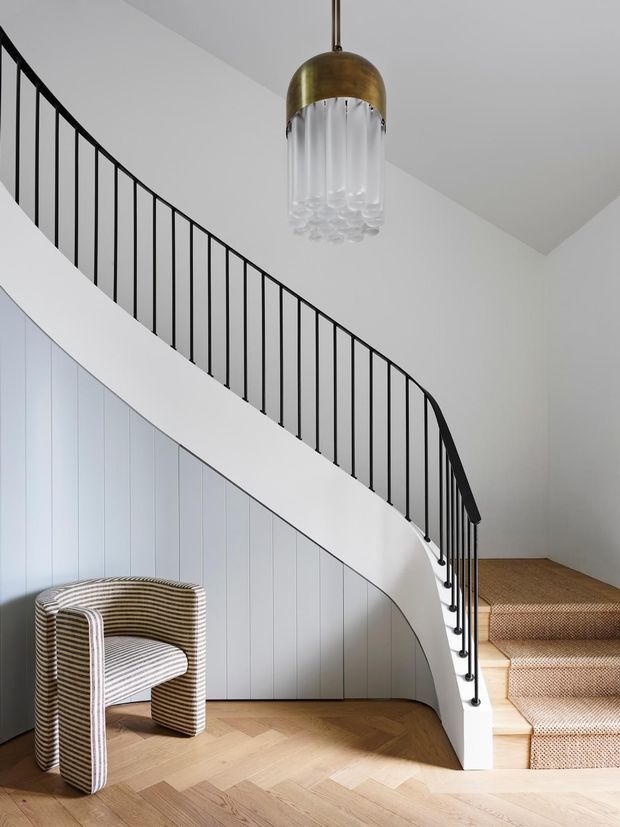 A refined interior stairway with a sweeping curved staircase, slim black metal balustrade, white vertical wall panelling, and warm herringbone timber flooring. The stairs are carpeted in a textured neutral beige, and a striped upholstered armchair sits beneath the stair. A sculptural pendant with a brass dome and cascading clear glass elements hangs overhead.