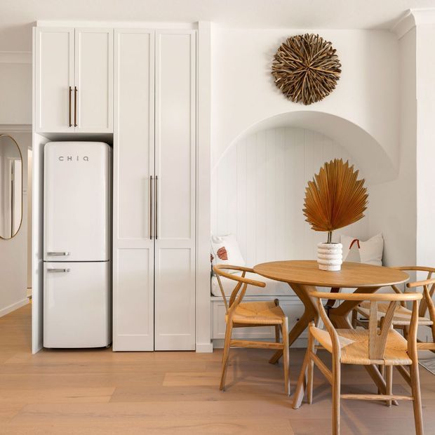 A warm, light-filled dining area adjoining a compact kitchen, featuring a round timber dining table, woven-seat timber chairs, a freestanding retro-style white refrigerator, and tall white paneled cabinetry. The room has pale timber flooring, white walls, an arched recessed feature with vertical paneling, and natural sculptural wall and table decor.