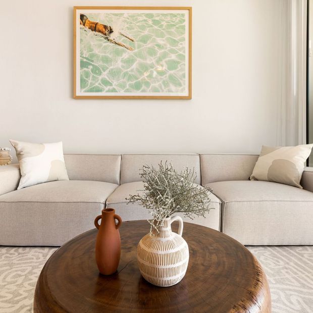 A softly styled living room with a large light-beige sectional sofa, neutral cushions, a round dark timber coffee table, ceramic vases with dried branches, a pale patterned rug, and a framed abstract artwork above the sofa. The room has light walls, sheer curtains, and a calm, refined neutral palette.