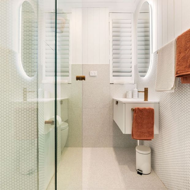 Bright compact bathroom featuring a frameless glass shower enclosure, white hexagonal wall tiles, pale speckled floor and wall tiles, white vertical wall panelling, plantation shutters, a wall-mounted white vanity, and warm rust-coloured towels. Brass and white bathroom accessories add subtle contrast.
