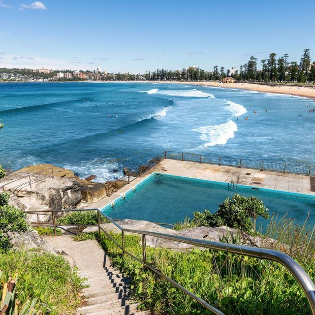 A bright coastal landscape viewed from a rocky headland, with a public ocean pool in the foreground, stairway and metal handrails descending toward the pool, and a sandy surf beach and urban shoreline across the bay.