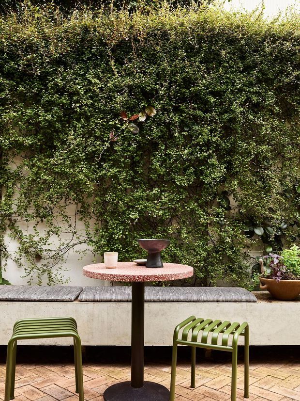 A refined outdoor courtyard seating area with a dense vertical green wall covering a rendered boundary wall. A round terrazzo-look table with a dark pedestal base is paired with two olive-green slatted chairs, while a built-in concrete bench with a grey cushion runs along the rear wall. Potted plants and patterned paving create a lush, intimate garden setting.