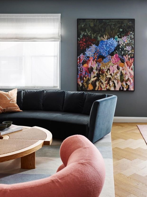 A stylish living room with a deep charcoal-grey wall, large colorful floral artwork, sheer Roman blinds, a black curved sofa, and a sculptural blush-pink lounge chair. A round wood coffee table with woven cane insets sits over a neutral rug, while pale herringbone timber flooring adds warmth.