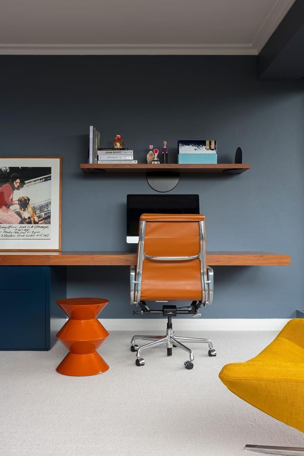 A stylish home office with a dark blue-grey feature wall, built-in dark cabinetry and a long timber desk. A vintage-style tan leather office chair sits centrally, with a floating timber shelf displaying books and small decorative objects. The room includes a bright orange sculptural side table, a partially visible mustard-yellow lounge chair, framed artwork, and light carpet. White ceiling cornices and polished metal chair details add contrast.