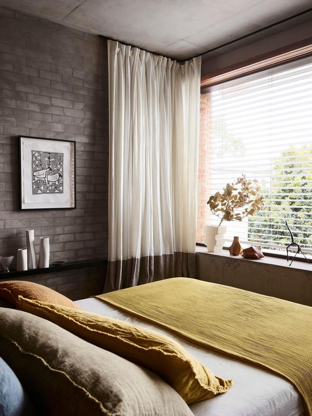 A warm, refined bedroom corner with a bed dressed in layered neutral and mustard-yellow textiles. The room features exposed grey brick walls, a concrete ceiling, a large window with horizontal blinds, full-height cream curtains with a darker lower band, and a dark built-in ledge displaying minimalist ceramics and a framed monochrome artwork. Natural light and earthy materials create a calm, tactile atmosphere.