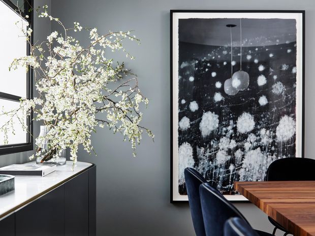 A refined dining area with a dark charcoal-grey wall, a large black-framed abstract artwork, and a substantial timber dining table with dark upholstered chairs. A black built-in sideboard with a pale stone countertop sits beneath a black-framed window, decorated with flowering branches. The composition has a moody, sophisticated contemporary feel with strong contrast between dark finishes, pale stone, white flowers, and warm timber.