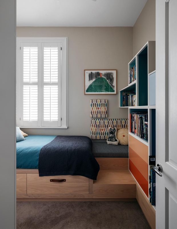 A compact, well-appointed bedroom viewed through an open white door. The room features a built-in timber platform bed with drawers, blue bedding, a patterned upholstered window seat, white plantation shutters, and a floor-to-ceiling built-in bookshelf with contrasting dark blue interiors and colourful drawers. The palette is warm and muted with playful geometric accents.