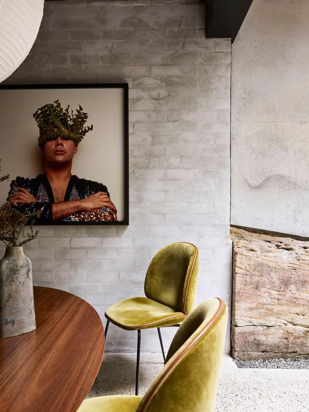 A moody dining area with a round dark timber table and two olive-green upholstered dining chairs. The space features white-painted exposed brick walls, a pale speckled floor, a rough stone fireplace or hearth edge, and a large framed portrait on the wall. A dark structural beam and sculptural pendant light add architectural character.