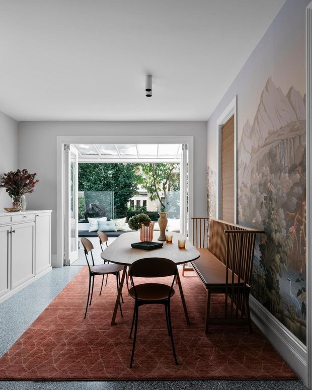 A refined dining room with a round dining table and dark modern chairs, positioned between a white built-in sideboard and a large scenic mural wall. A glazed opening leads to a bright outdoor courtyard or terrace with additional seating. The room has pale grey walls, a white ceiling, a muted red patterned rug, timber accents, and abundant natural light.