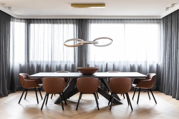 A refined dining room centered on a long rectangular dining table with sculptural dark legs and six brown upholstered dining chairs. Floor-to-ceiling sheer and charcoal-grey curtains frame expansive glazing, while a contemporary sculptural pendant hangs above the table. The room has a pale herringbone timber floor, white ceiling, and a minimalist, polished atmosphere.