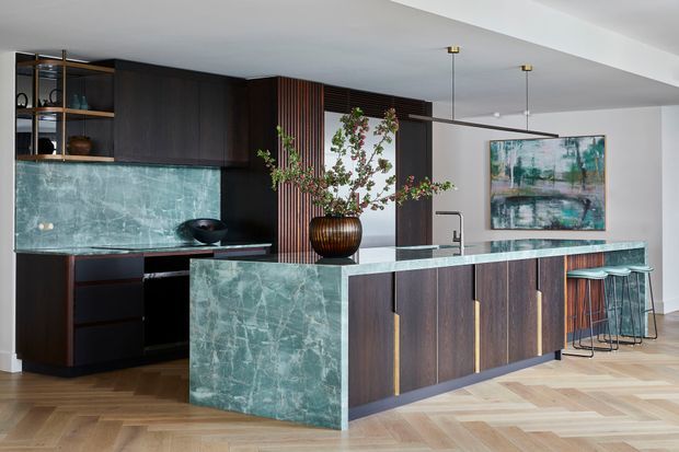 Contemporary open-plan kitchen featuring dark timber cabinetry, a large island with a dramatic green veined stone waterfall benchtop, integrated appliances, open shelving, a vertical timber slat feature, and bar seating. Pale herringbone timber flooring and pendant lights add warmth and contrast.