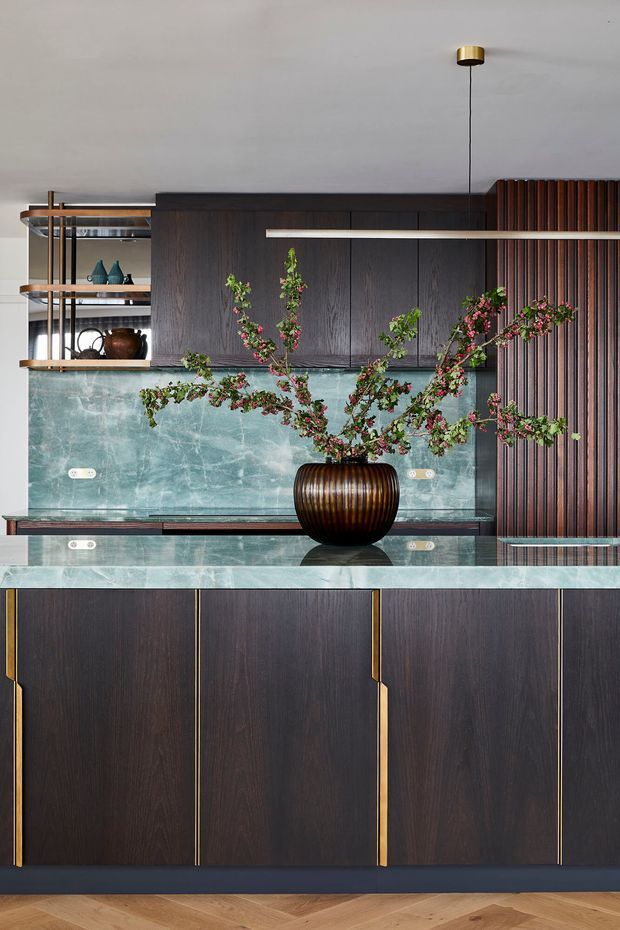 A refined contemporary kitchen featuring dark timber cabinetry with slim brass-toned pulls, a pale green veined stone benchtop and splashback, open timber-and-metal shelving, and a vertical timber battens feature wall. A sculptural dark vase with flowering branches forms a central decorative feature, while a linear pendant light hangs above.
